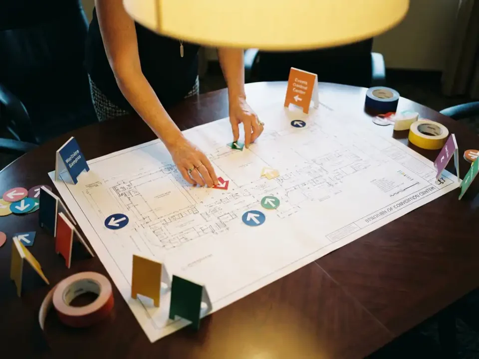 Handen van eventplanner die pijlborden en vloermarkeringen rangschikt op gedetailleerde plattegrond van evenementenlocatie